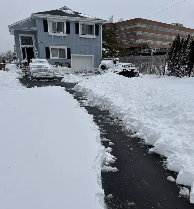 A path cleared by shovel through deep snow leading up to a blueish grey house with white windows and doors and black shutters