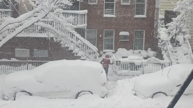 A person holding a shovel in a blizzard surrounded by snow-covered cars