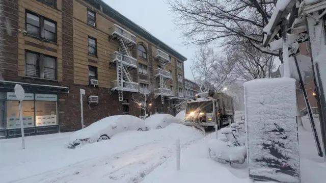 A New York street covered in snow. Cars parked on the side are completely covered in a layer of snow. There is a snow plough going through the road, trying to clear it