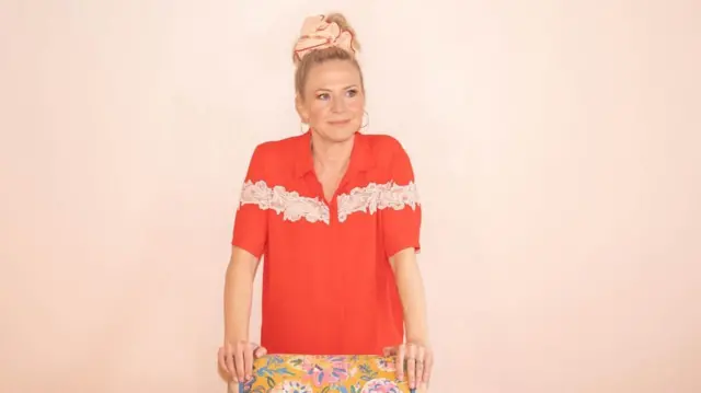 Actress Kellie Bright in a bright red shirt standing behind a floral patterned chair against a pink background.