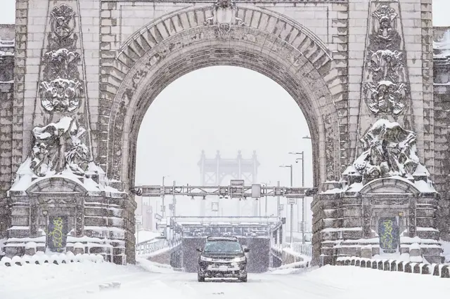 A car coming through a large arch at the end of a bridge during a blizzard
