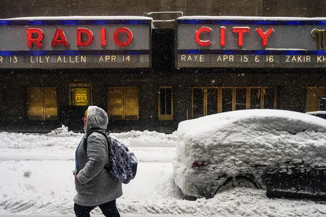 A person in grey hooded coat and backpack walks through the snow outside Radio City Music Hall