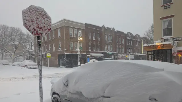 A frozen stop sign on a Brooklyn street, with a car covered in snow.