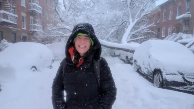 A woman in a large winter coat stands in front of a fallen tree on a snow-covered Brooklyn street