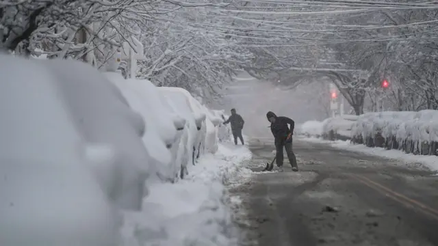 People stand on a road, which has been gritted, shovelling snow away from the parked cars. There is a very thick layer of snow on top of the cars, meaning we cannot see anything but the tips of their wheels.