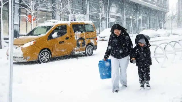 A mother and son walk along covered in snow, beside a yellow New York city taxi and a