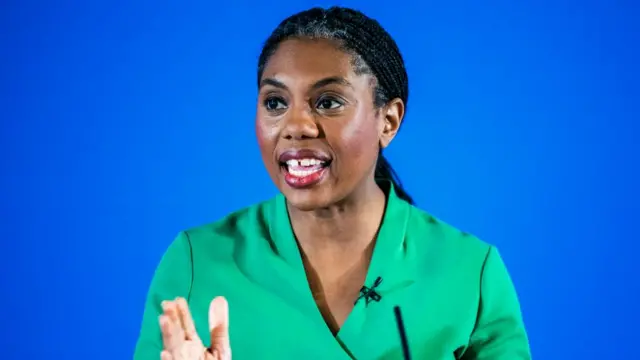 Conservative leader Kemi Badenoch wearing a green dress and standing in front of a blue background