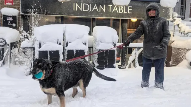 A dog wagging its tail in the snow with a blue ball in its mouth. Its owner is stood next to it, holding onto the lead and smiling