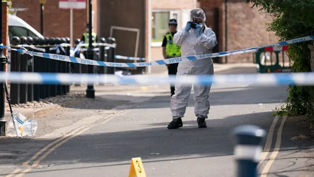 View of the crime scene. Blue police tape hangs across the road and a forensic officer in a white protective suit takes photographs