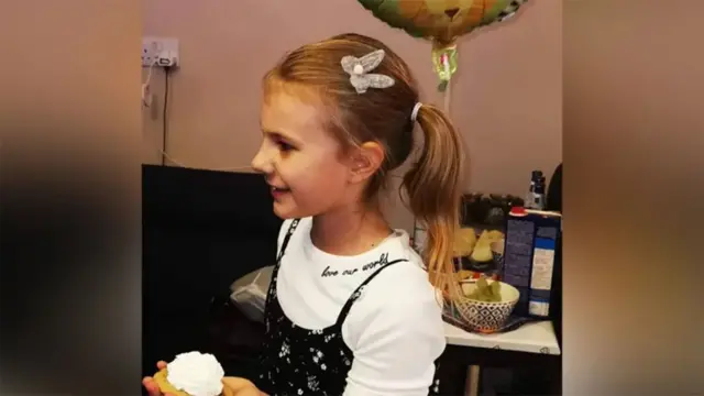 Lilia holding a cake. She has blond hair in a ponytail and is wearing a white top and black patterned dress
