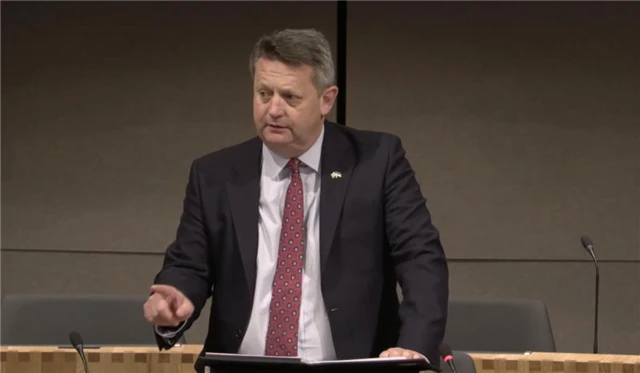 A man stood up talking. He has a black jacket on and a light pink shirt with a red tie. He has short grey hair.