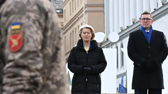 Usula von der Leyen stood next to Estonian PM Kritsen Michal. They're both dressed in black. A Ukranian soldier in military camouflage uniform is walking past them, blurred in the foreground