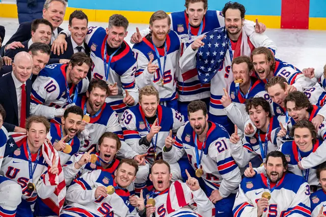 The US men's hockey team in white, red and blue jerseys appears to shout in celebration while holding up one finger and the hand holding their gold medals