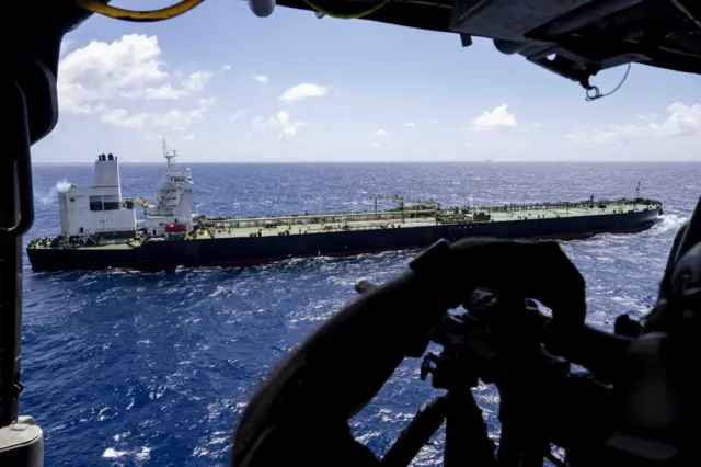 A picture of a tanker sailing in the ocean taken from a military helicopter, you can see a soldier aiming a rifle in the foreground
