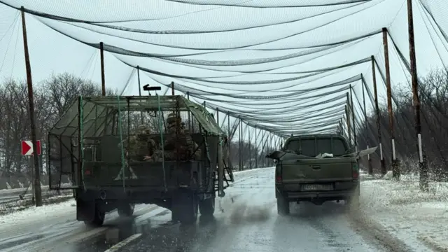 A vehicle carrying Ukrainian soldiers is covered in netting next to a truck. Poles have been put up along the motorway which have fishing nets drapped over them