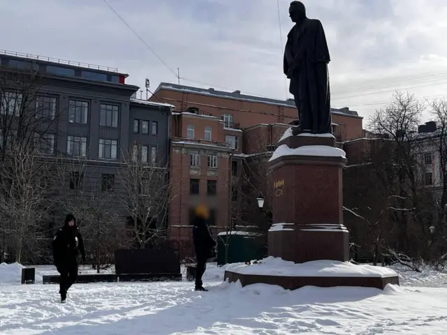 A statue in the snow. Two people stand by it, their faces blurred, paying tribute