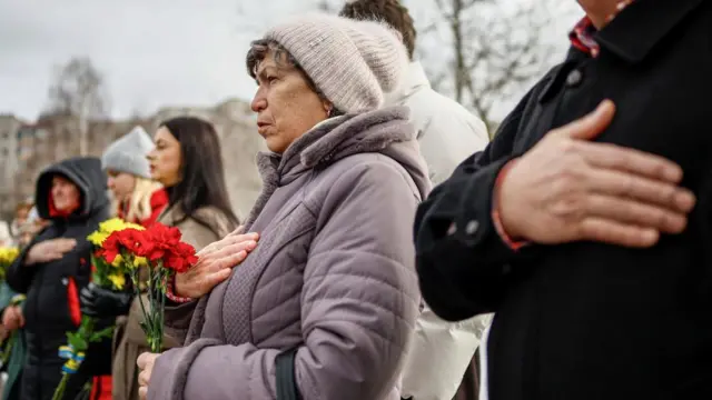A row of people hold flowers and place a hand on their chests during a memorial service in Bucha, Ukraine.