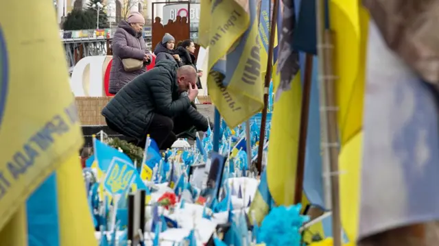 A man visits a makeshift memorial for Ukrainian soldiers in Kyiv. He has his head in his hands as he crouches down to the flags on the ground.