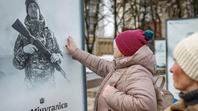 A woman in winter clothes holds a hand towards a black and white photo of a Ukrainian soldier who died in the war