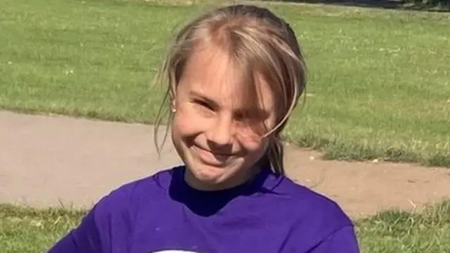 Lilia Valutyte she has blond hair and is wearing a purple top and is in a park with grass behind her