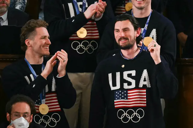 A beared man in a USA sweater holds up an Olympic gold medal