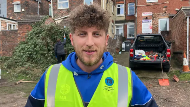A close-up of a man with curly brown hair looking at the camera. He wears a blue hoodie and yellow hi-viz jacket with a blue TS logo on it.