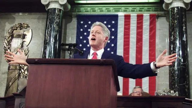 Bill Clinton holds his arms out at a podium in front of flag