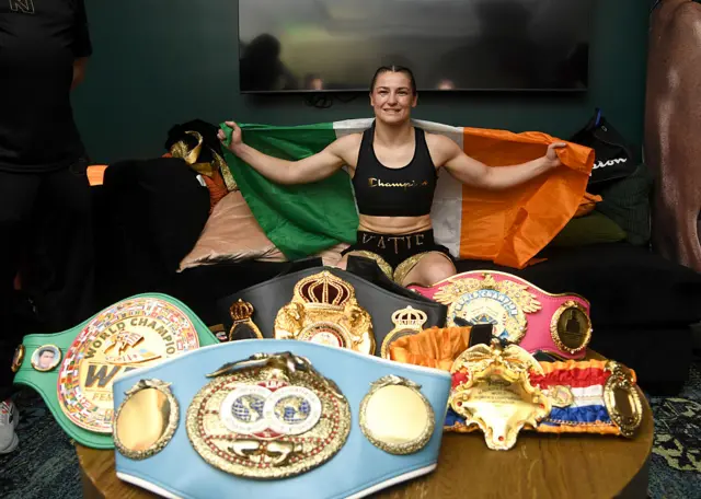 Katie Taylor sits on a sofa and holds the Irish flag behind her back with her light-welterweight titles on a table in front of her