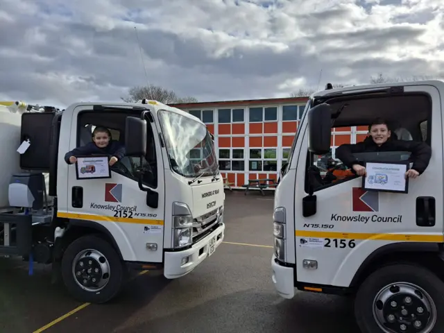 Two bin wagons parked facing each other in a car park, with a boy aged about 10 sat in each cab. THe boys look out of the winow and hold certificates.