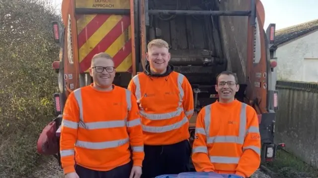 Three men in bright orange visibility jackets stand in front of a bin lorry.