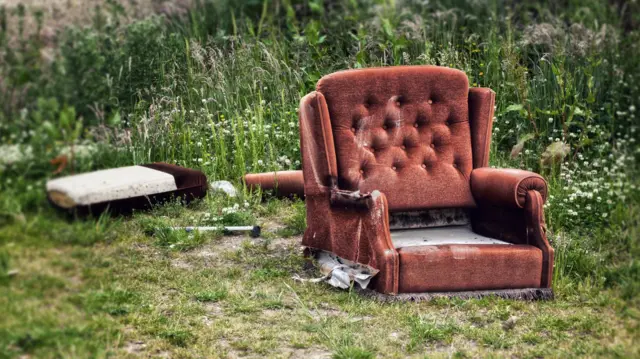 A old sofa has been dumped on a patch of grass. There are white wild flowers in the background.