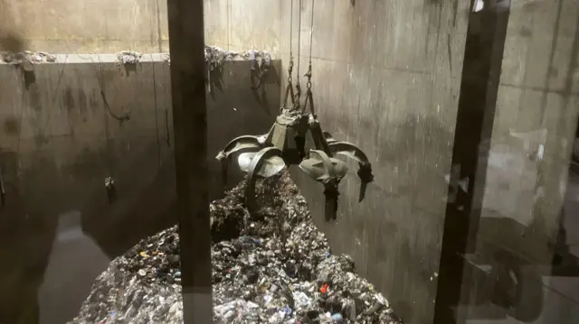 A large indoor waste facility where piles of household rubbish sit on the floor, with cranes above ready to grab rubbish lift material into an incinerator.