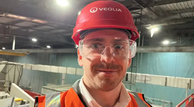Oliver Peat, a man in a recycling facility wearing an orange hi-vis vest, protective glasses and a red hard hat