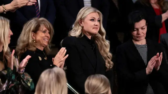 A blonde woman dressed in black stands while those around her applaud