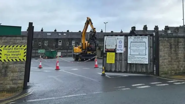 A recycling centre in Ilkley, West Yorkshire, showing a digger and some metal shipping containers within the site's wooden gates.