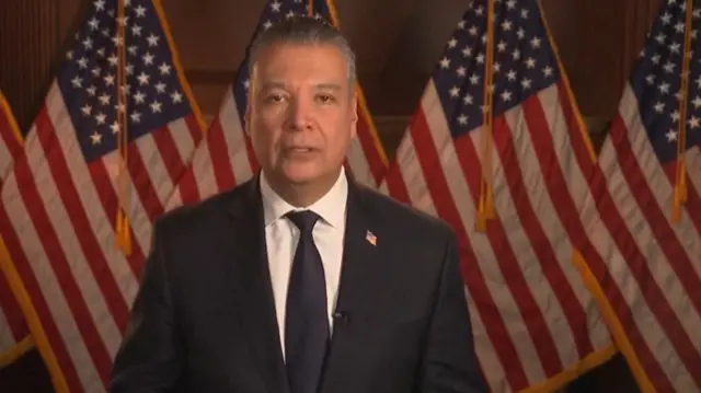 Alex Padilla wearing a black suit over a white shirt and black tie. There are American flags behind him.