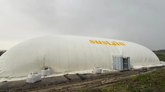 A large white inflated dome made from tent-like material. It is rounded, tall and in an oblong shape with an entryway. The word Sustain is printed on it in yellow. It sits on a muddy field under a grey sky.