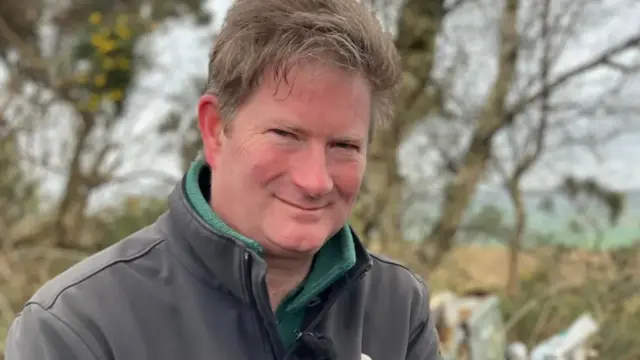 A man in a grey fleece in standing in front of a hedegrow in rural Devon. There is fly-tipped material in the background including a black bin bag