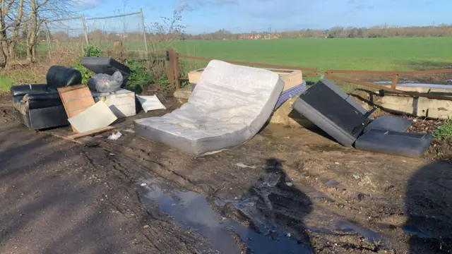 Fly-tipping at a gate next to a field. The waste includes a sofa, a mattress and other rubbish. A shadow of the person taking the photo can also be seen.