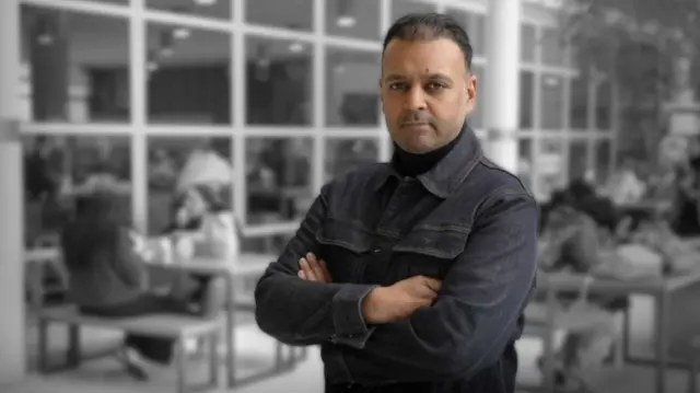 A man poses in a cafe, looking into the camera with his arms folded. He is wearing a turtle-neck black jumper and a dark jacket
