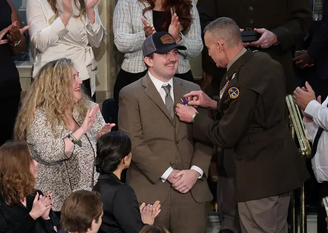 A military official pins a medal on the lapel of a man in a brown suit and baseball hat