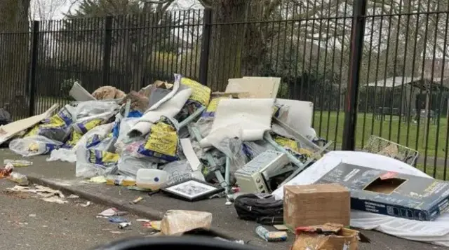 A large amount of fly-tipped rubbish on a street next to a metal fence with a park in the background