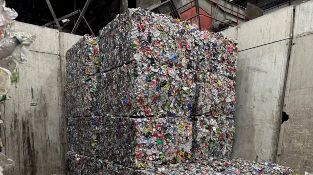 Stacks of tightly compressed bales made from crushed aluminium cans are piled high inside an industrial waste‑processing facility. The bales form large rectangular blocks against tall concrete walls, with loose debris scattered across the floor.