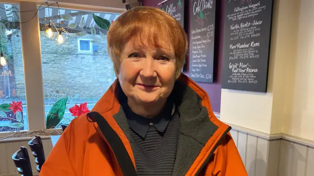 A woman in an orange coat stands in a cafe. Chairs and tables can be seen behind her