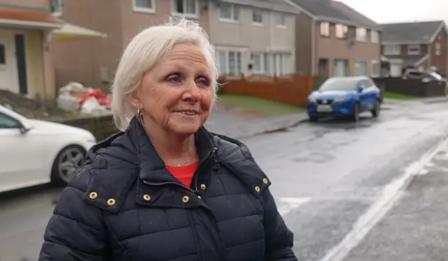 Anne Crimmage, a woman wih white hair wearing a red jumper and black coat, standing on a street in Wales.