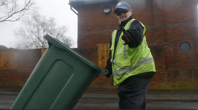 A man in a yellow h-vis vest wheels a bin down the street
