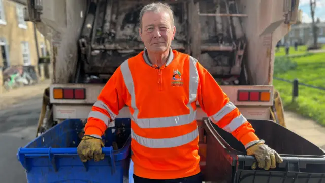 Chris Lively looking into camera. He has short hair and is wearing an orange council jumper, and yellow gloves. He is standing in between two bins, which he is holding, and in front of the back of a bin lorry.
