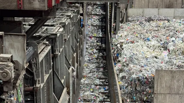 A industrial waste‑processing facility filled with large amounts of mixed rubbish, primarily plastic. In the centre of the scene, a long conveyor belt runs vertically through the frame, carrying scattered plastic debris along its surface. To the left, there is a heavy metal structure made up of panels, machinery components, and support beams
