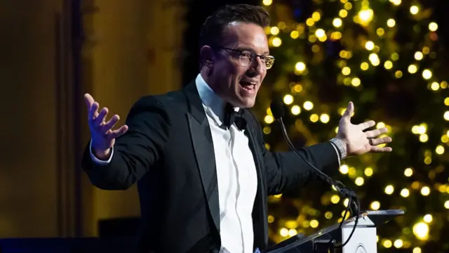 Benny Johnson speaks at a microphone while wearing a tuxedo