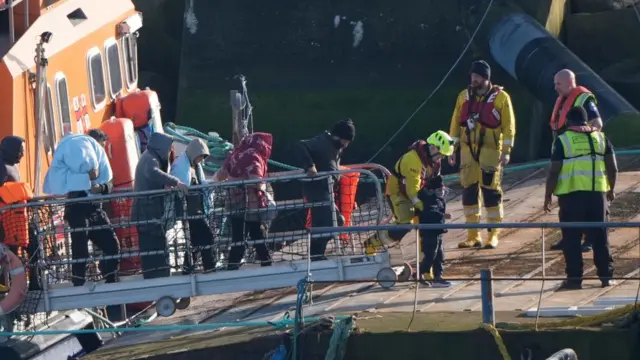 People walking a gangway from the ship to the harbour. Many wear blankets around them. Four emergency personnel stand on the harbour waiting for the migrants. One child is being helped by the worker